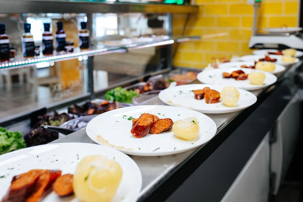 Slices of traditional Hungarian smoked sausage and a pickled apple pepper served on white plates lined up on a restaurant counter.
