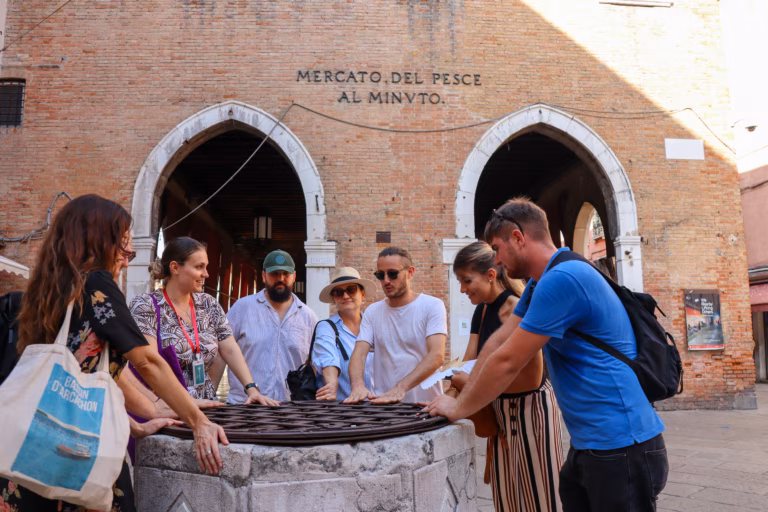 A small group gathers around an old stone wellhead outside the "Mercato del Pesce al Minuto" fish market entrance in Venice.